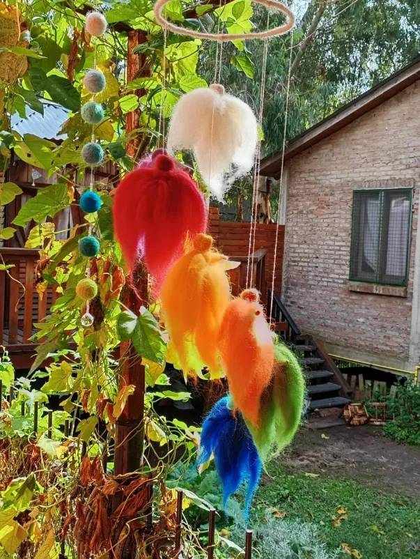 Móvil de figuras de lana afieltrada en colores del arcoíris —blanco, rojo, naranja, verde y azul— colgando de un aro de madera al aire libre, entre plantas.