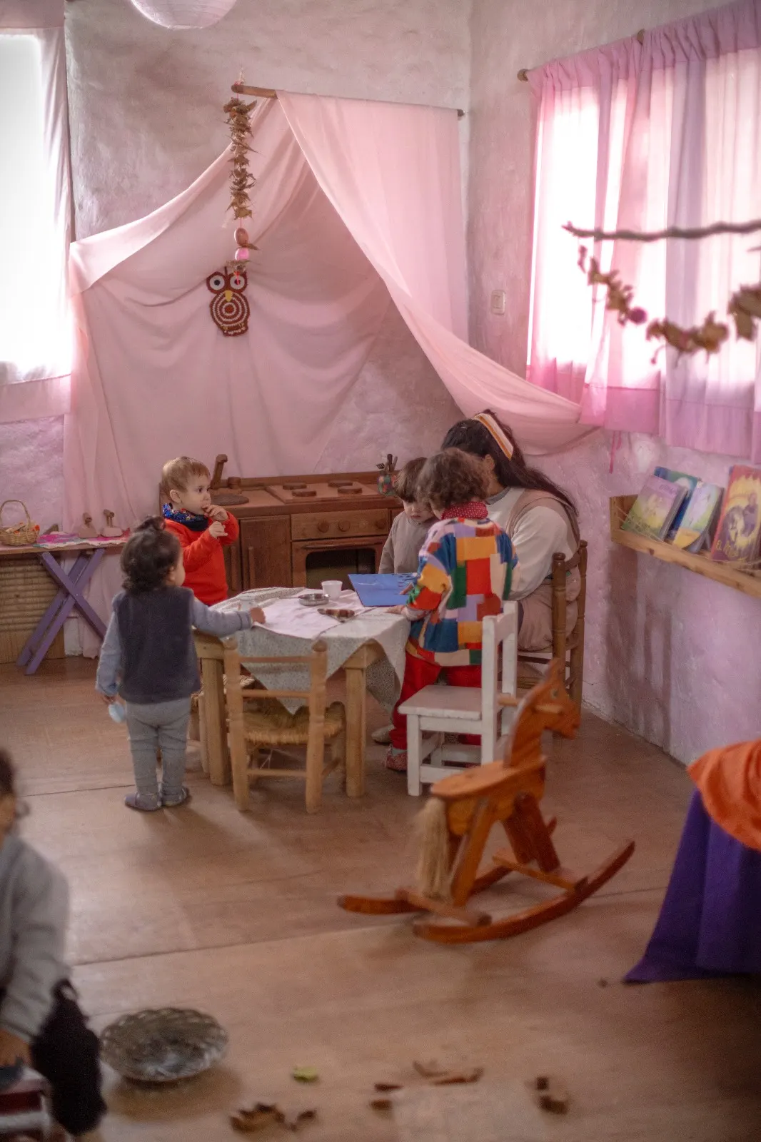 Maestra y niños pequeños jugando junto a una cocinita de madera en una sala cálida del jardín